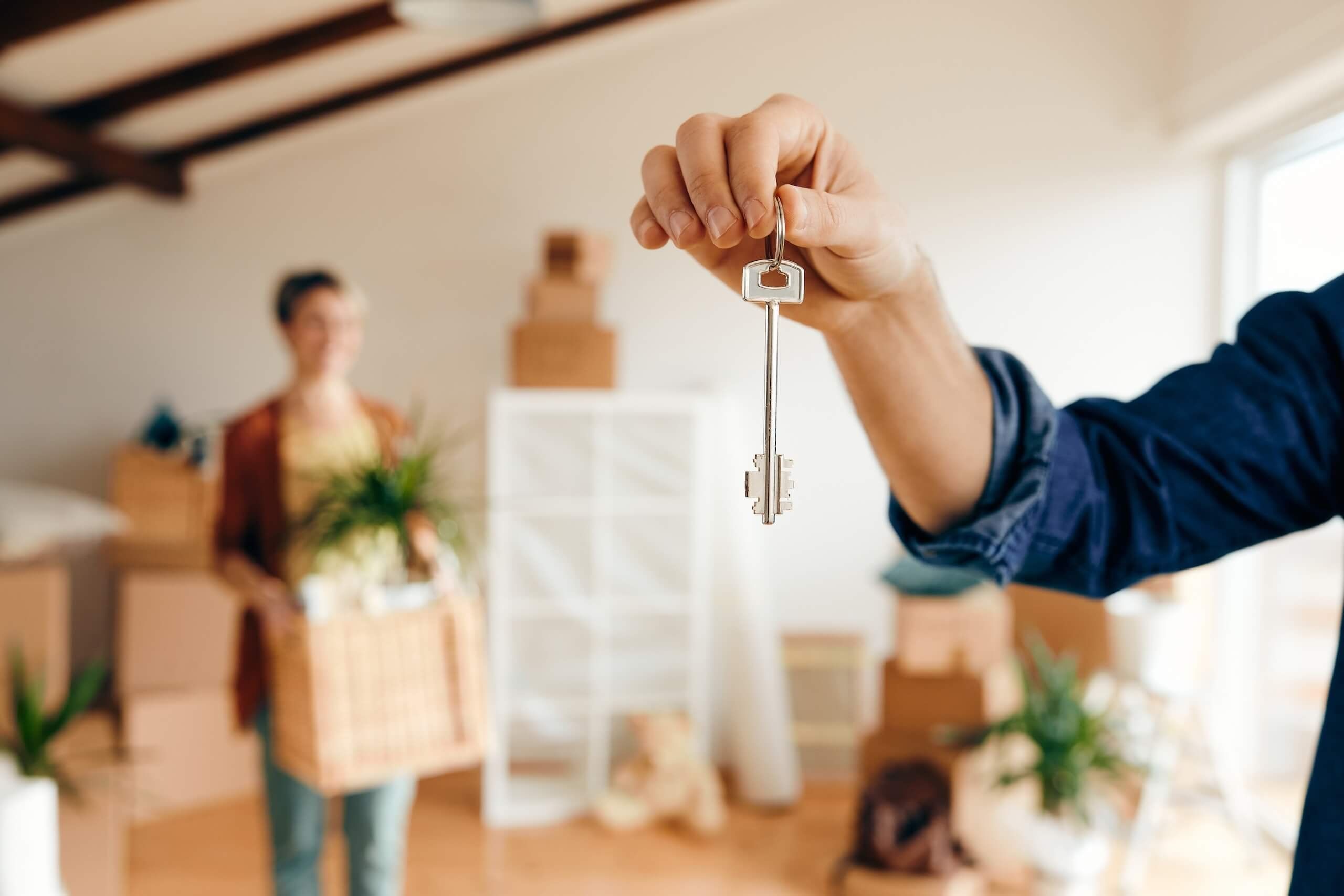 Hand holding up a house key while a woman carries a box into a new home