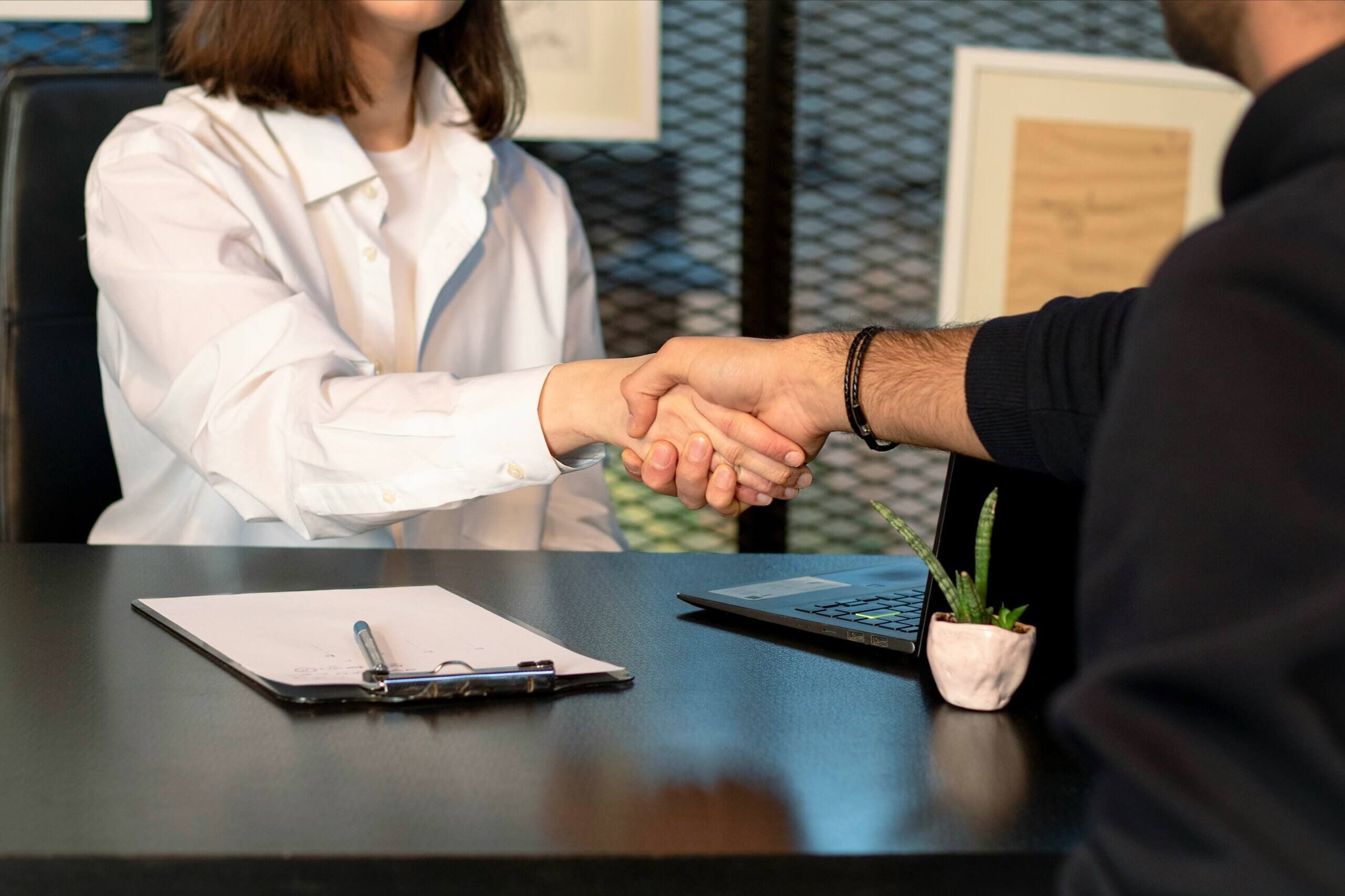 Two people shaking hands across a desk with a clipboard and a laptop