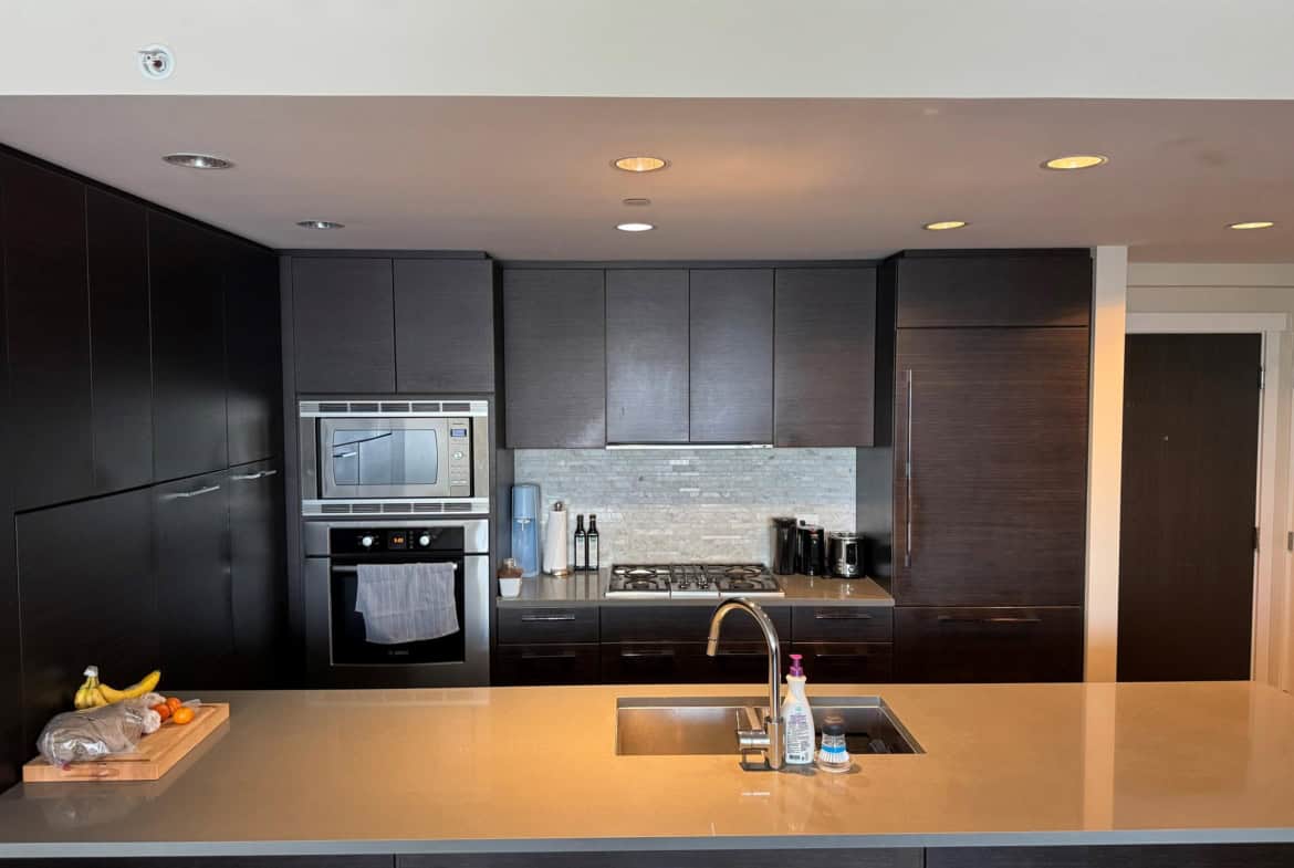 Modern kitchen with dark wood cabinetry, built-in oven and microwave, beige countertop, and stainless steel appliances including a kettle and coffee maker, ideal for Vancouver Island homebuyers.