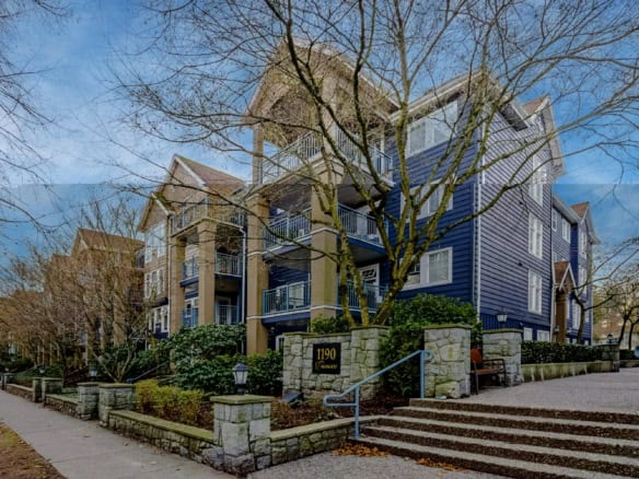 Modern multi-family residential building with blue exterior siding, balconies, and stone accents, surrounded by trees and landscaped gardens in a neighbourhood with sidewalks and street lighting.