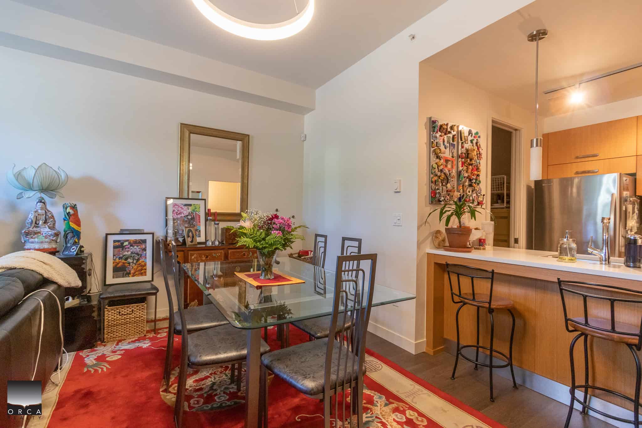 Bright dining area with glass table, decorative plants, and colorful artwork, adjacent to modern kitchen with stainless steel appliances, ideal for Vancouver Island real estate homes.