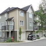 Modern multi-family townhouse with beige and gray siding, featuring private balconies and attached garages, located in a peaceful residential community.
