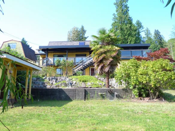 Spacious multi-level yellow house with large deck, surrounded by lush greenery, trees, and landscaped garden, set in a serene residential neighbourhood in British Columbia.