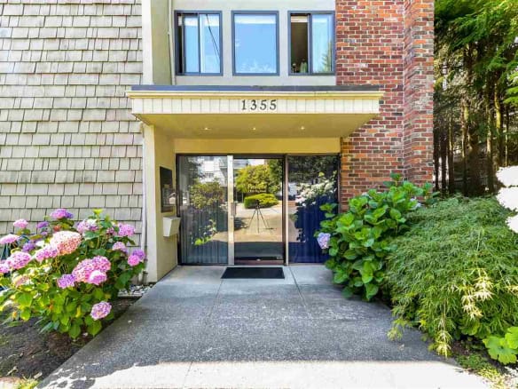 Modern apartment building entrance in Vancouver with lush greenery and vibrant hydrangea flowers, featuring a glass door and brick accents, ideal for real estate listings.