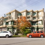 Modern multifamily apartment building in urban Vancouver, BC with green and beige exterior, balconies, parking, and autumn tree in front.