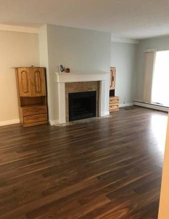 Cosy living room with wooden flooring, a fireplace with a white mantel, and built-in wooden cabinets on either side, illuminated by a large window with white curtains.