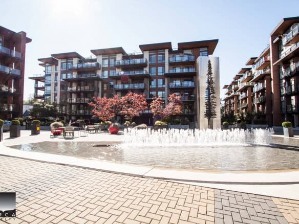 Modern multi-story residential buildings with balconies and glass railings in a landscaped setting featuring a central fountain, outdoor seating, and pink blossoming trees in a contemporary urban complex.