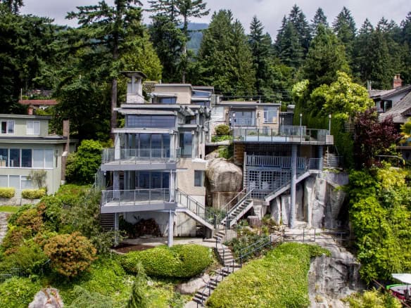 Modern hillside luxury home with expansive glass windows, surrounded by lush greenery and mature trees, offering stunning views, prime real estate, and upscale living in a vibrant community.