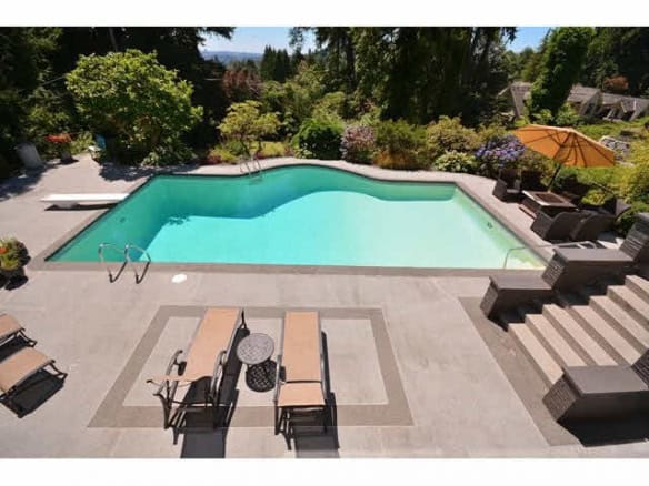 Aerial view of a modern backyard swimming pool with surrounding patio furniture, lush greenery, vibrant trees, and an umbrella in a private residential setting.