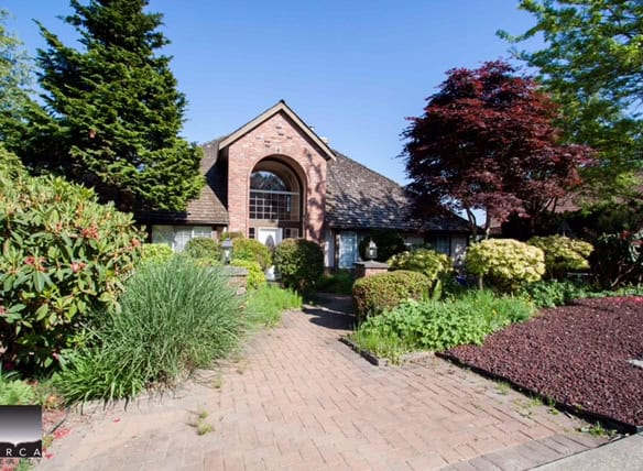 Lush landscaped garden leading to a beautiful brick house with large arched window, surrounded by vibrant trees and shrubs, showcasing Orca Realty Inc.'s prime properties in Ontario.