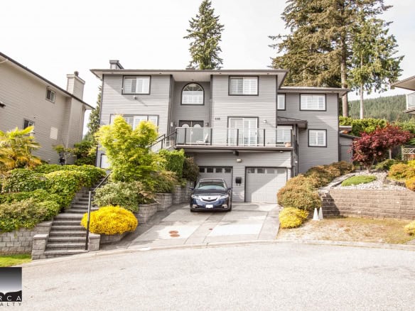 Modern multi-level grey residential home with landscaped front yard, driveway, and mountain backdrop, offered by Orca Realty Inc. in BC.