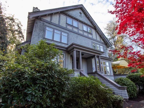 Luxurious Victorian-style house with grey wooden exterior, large windows, surrounded by lush greenery and vibrant autumn foliage, available through Orca Realty Inc. in Vancouver.