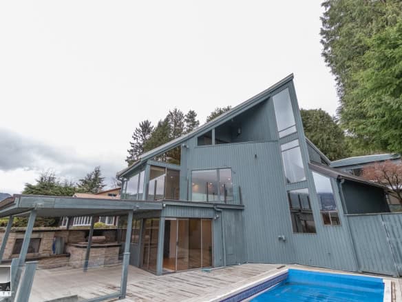 Contemporary modern house with angular roofs, large glass windows, and a backyard with a pool, located in a scenic area with trees and mountains, ideal for real estate investments.