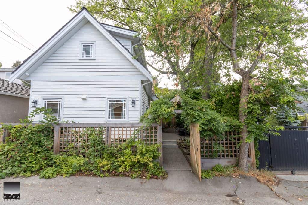 Charming white family home near outdoor greenery, featuring a small fenced yard and mature trees, ideal for neighbourhood real estate in Victoria, BC.