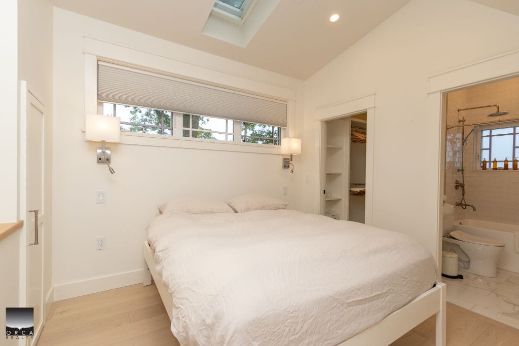 Modern minimalist bedroom with neutral tones, adjacent bathroom, and skylight for natural light, showcasing upscale interior design for Vancouver Island real estate.