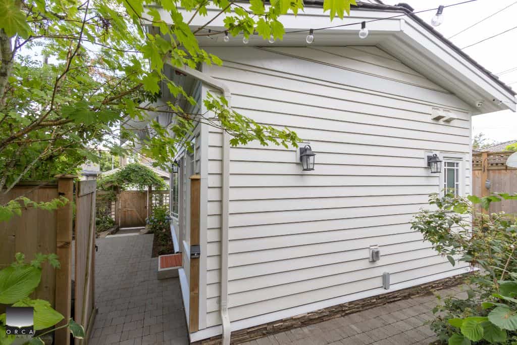 White exterior house with outdoor lighting located in a backyard, featuring a pathway surrounded by greenery, fence, and trees, showcasing residential property in Vancouver.