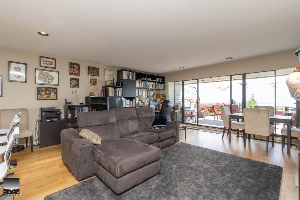 Modern living room in a Vancouver-area condo with open floor plan, featuring a brown sectional sofa, artwork on the wall, a home office setup, and large sliding glass doors leading to a balcony.
