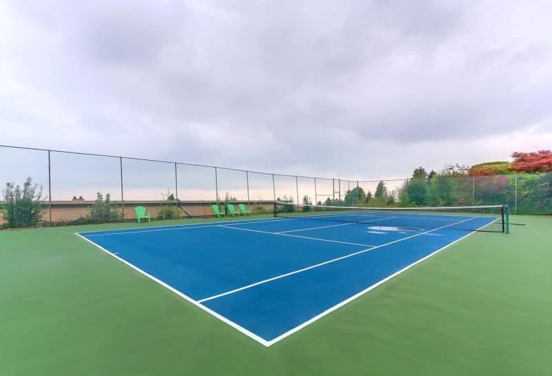 Tennis court at Orca Realty Inc. property with a vibrant blue playing surface, surrounded by greenery and a safety net, showcasing premium real estate amenities and outdoor lifestyle opportunities.
