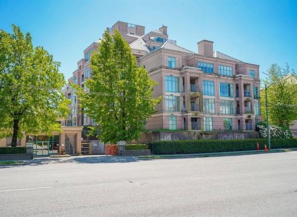 Luxurious multi-story residential building with modern architecture and large glass windows in a vibrant urban setting, surrounded by lush green trees and a clear blue sky.