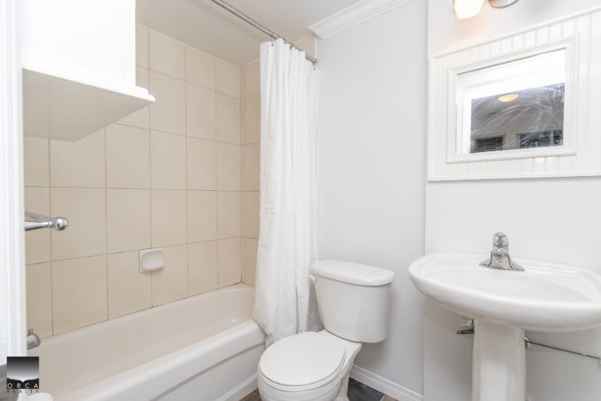 Bright bathroom with bathtub, toilet, and pedestal sink in a modern home. Featuring white tiles, fresh paint, and natural light from a small window, ideal for comfortable living in Vancouver.