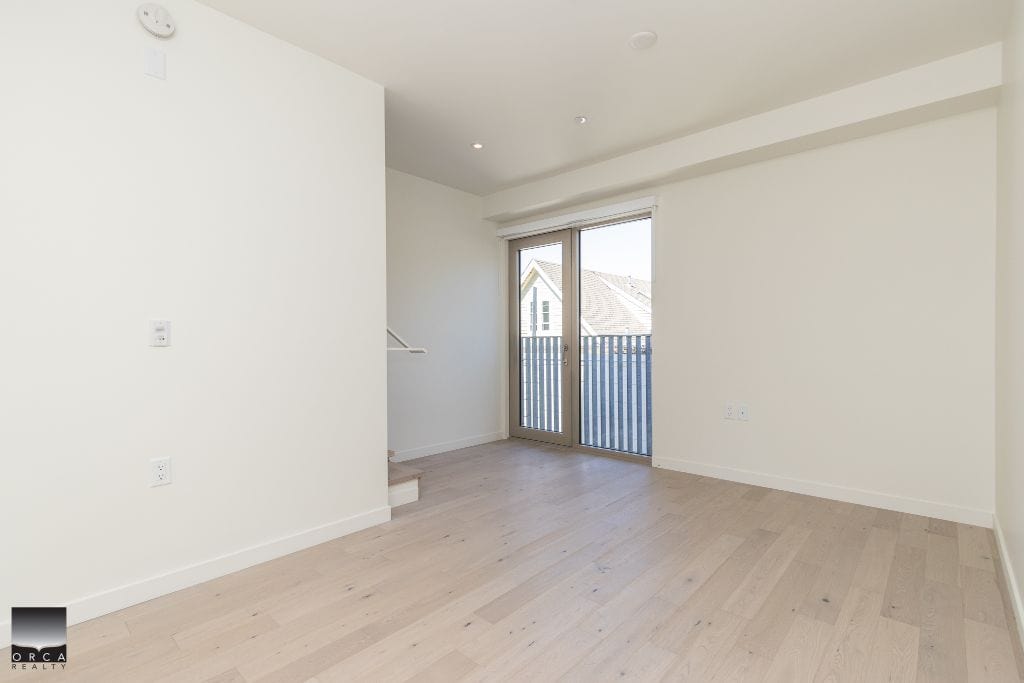 Bright, empty living room with large glass door leading to balcony, white walls, light wood flooring, modern interior, ideal for homebuyers looking for Vancouver Island real estate.