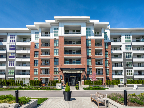 Modern multi-story apartment building with red brick and white accents, featuring balconies, landscaped courtyard, and clear blue sky, representing premium residential real estate and property management services in Vancouver.