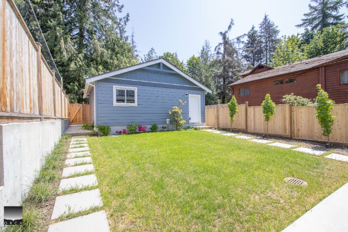 Bright blue backyard cottage with white trim, lush green lawn, and concrete stepping stones, surrounded by a wooden privacy fence, perfect for Vancouver Island property, offered by Orca Realty Inc.