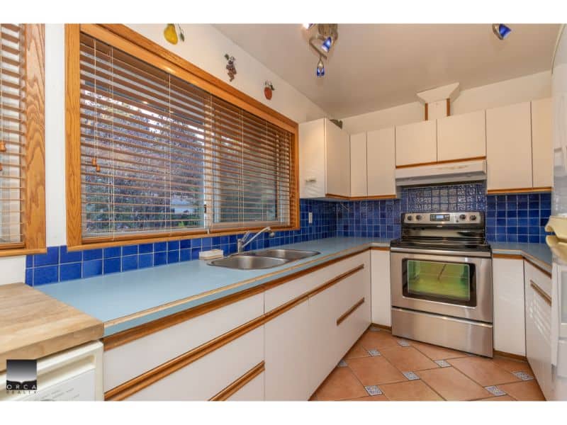 Modern kitchen with white cabinets, blue tiled backsplash, stainless steel oven, and large window with wooden blinds. Perfect for homebuyers seeking functional and stylish kitchen spaces.
