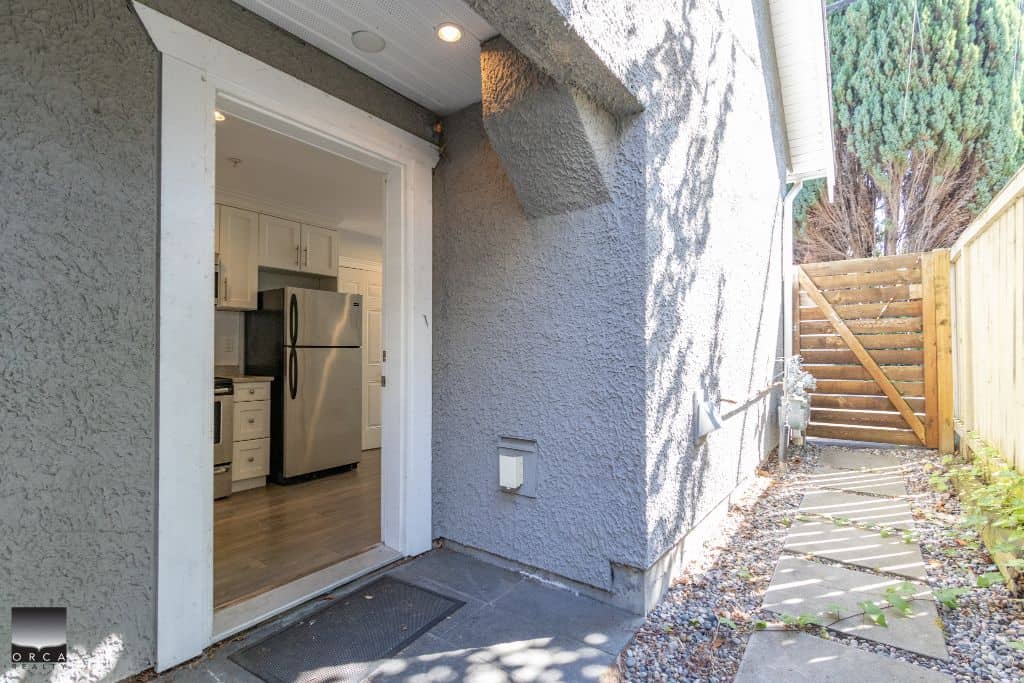 Modern kitchen seen from backyard door, stainless steel appliances, white cabinets, outdoor space with stone pathway and wooden gate, ideal for Vancouver real estate.
