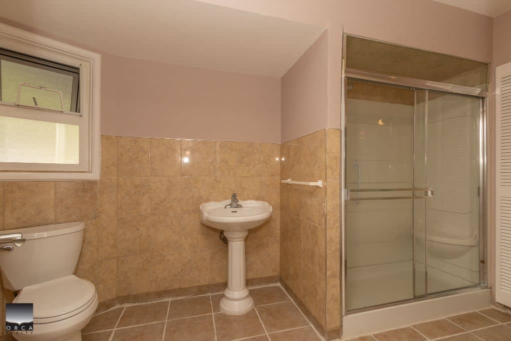 Modern bathroom with shower, toilet, and pedestal sink in a renovated home. Featuring beige tile accents, good natural light, and updated fixtures. Perfect for comfortable living.