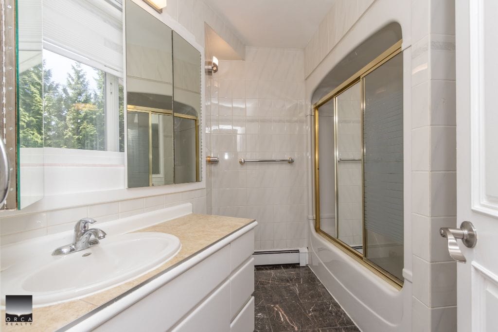 Bright clean bathroom with a white vanity, beige countertop, large mirrored cabinet, and a window with natural light. Features a bathtub with sliding glass doors and modern fixtures, perfect for Vancouver Island homes.