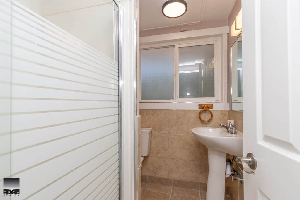 Bright small bathroom with shower, toilet, and sink, featuring neutral tile, a frosted window for privacy, and modern fixtures, ideal for Vancouver Island and BC real estate listings.