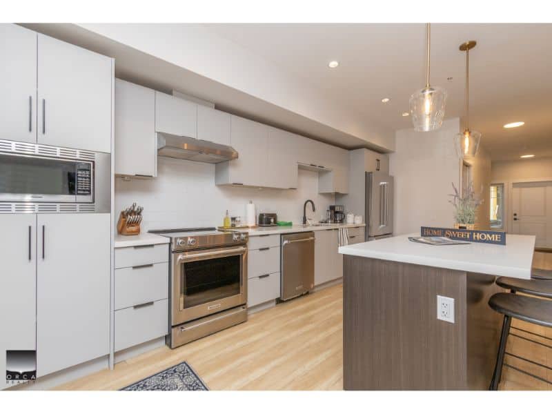 Modern white kitchen with stainless steel appliances and a spacious island, perfect for family living and entertaining in Victoria BC. Orca Realty Inc. specializes in Vancouver Island real estate.