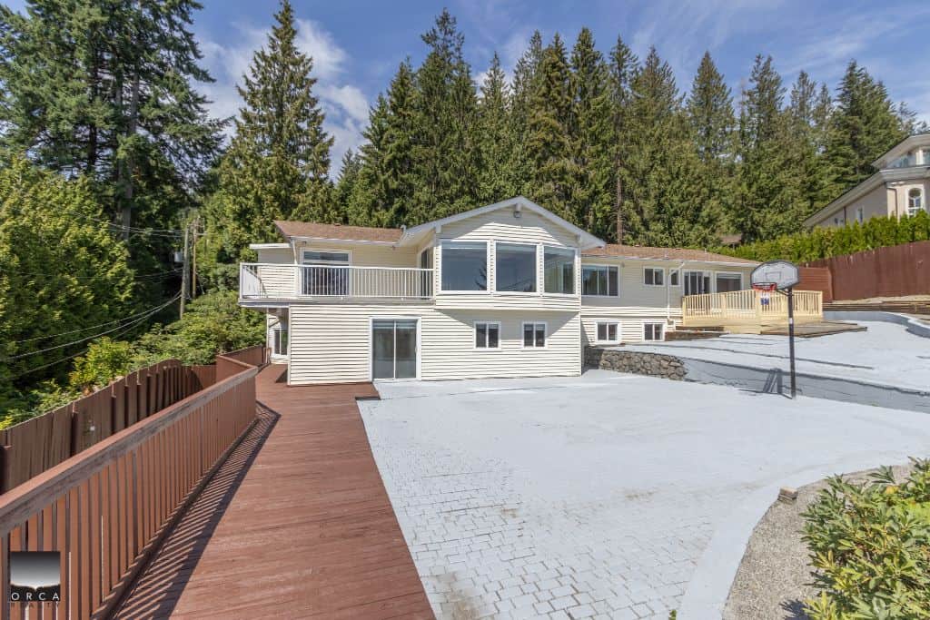 Spacious backyard with wooden deck and basketball hoop at a family-friendly home, featuring lush greenery and a scenic forest background. Perfect for outdoor living and entertaining.