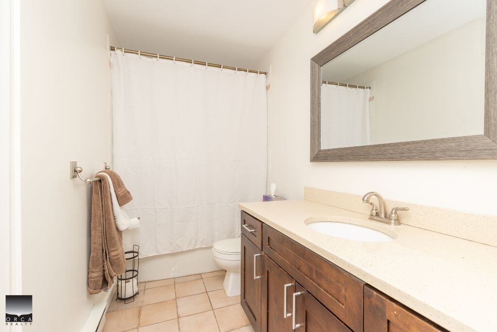 Bright bathroom with white walls, beige tile flooring, and a vanity with dark wood cabinets and a large mirror. Features a shower with a white curtain and a modern fixture for a comfortable, functional space.