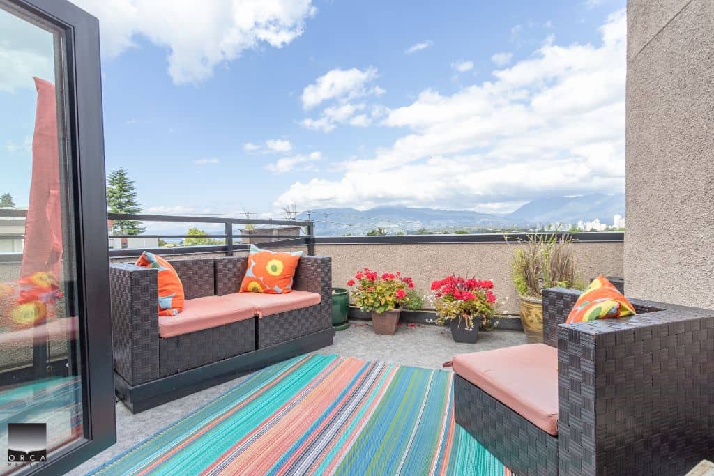 Balcony outdoor patio with colorful striped rug, wicker furniture with pink cushions, and vibrant potted flowers overlooking a scenic mountain view, perfect for relaxing or entertaining.