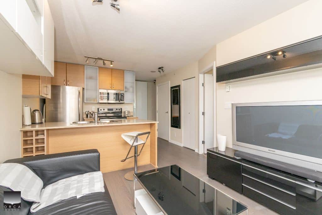 Modern open-concept living room and kitchen in a Vancouver condo, featuring a sleek black sofa, large flat-screen TV, and stylish kitchen with stainless steel appliances.