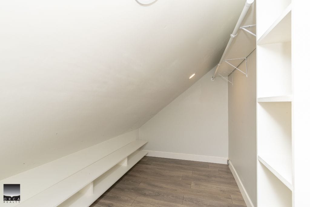 Spacious walk-in closet with white shelving and hanging rods, under sloped ceiling, featuring modern grey laminate flooring. Perfect for organized storage in a contemporary home.