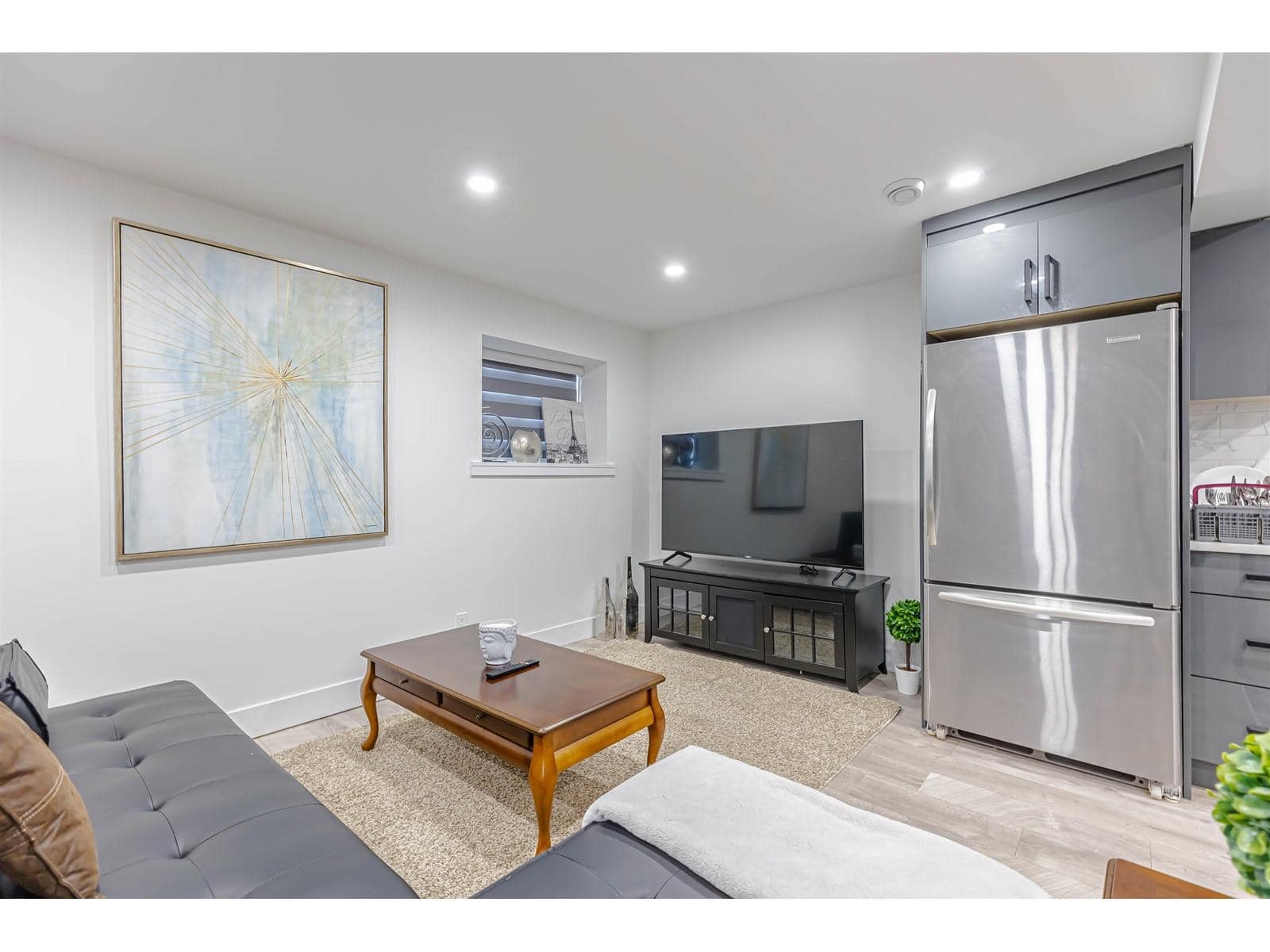 Warm modern basement living room with grey sofa, wooden coffee table, large flat-screen TV on black media console, stainless steel refrigerator, artwork, and minimalist decor.