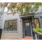 Contemporary grey townhouse exterior with modern black door, surrounded by lush greenery and trees, featuring large windows, ideal for Vancouver real estate listings and property sales.