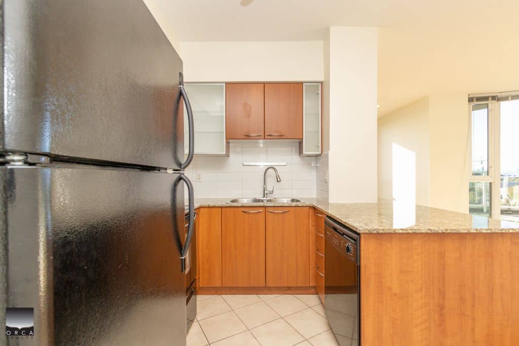 Modern kitchen with black refrigerator, wooden cabinetry, granite countertops, and stainless steel sink in a bright, open living space.