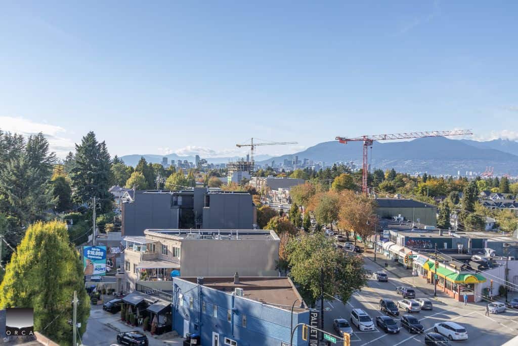 Modern urban neighbourhood with mixed commercial and residential buildings, construction cranes, and mountain views in the background.