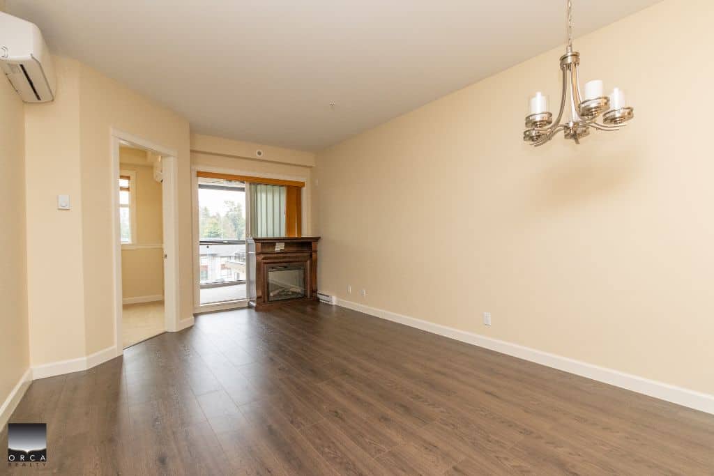 Spacious living room in a modern apartment with hardwood flooring, large window with balcony access, and a cozy fireplace, ideal for comfortable urban living in Vancouver, BC.