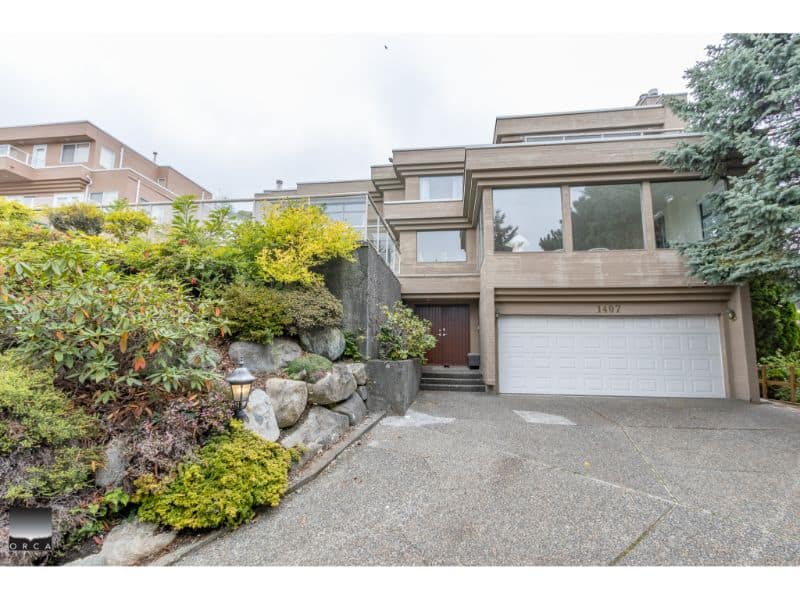 Modern multi-level grey residence with large windows and attached garage in Vancouver BC, featuring landscaped front yard with rocks and greenery, accessible driveway, and situated in a desirable neighborhood.