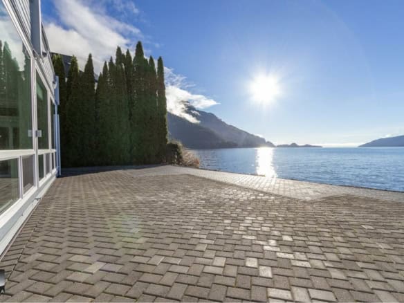 Stunning waterfront property with a paved patio overlooking a serene lake, mountain backdrop, and lush trees, perfect for enjoying Vancouver Island's natural beauty.