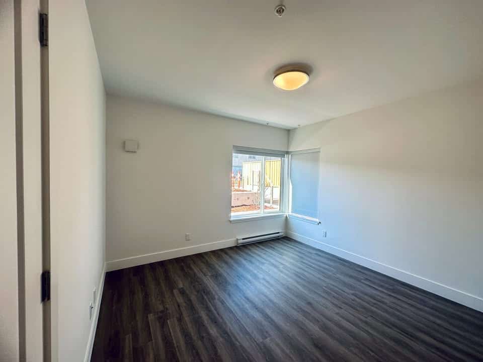 Modern vacant living room with large window, white walls, and dark hardwood flooring, ideal for first-time homebuyers and real estate investors in British Columbia.