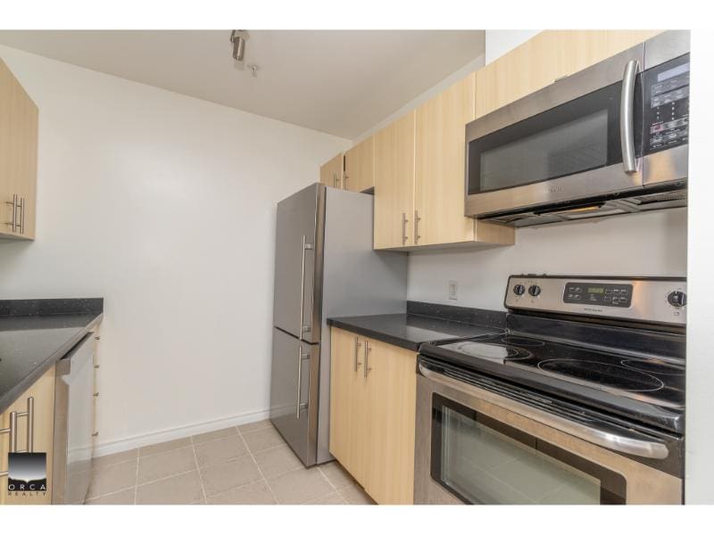 Bright modern kitchen featuring stainless steel appliances including a refrigerator, microwave, and oven with black countertops and light wood cabinetry, ideal for Vancouver Island real estate.