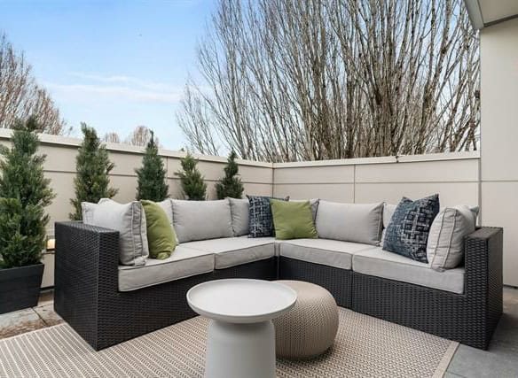 Elegant outdoor patio seating area with modern wicker sectional, plush cushions, potted evergreens, and minimalist side table, perfect for urban real estate outdoor living spaces.