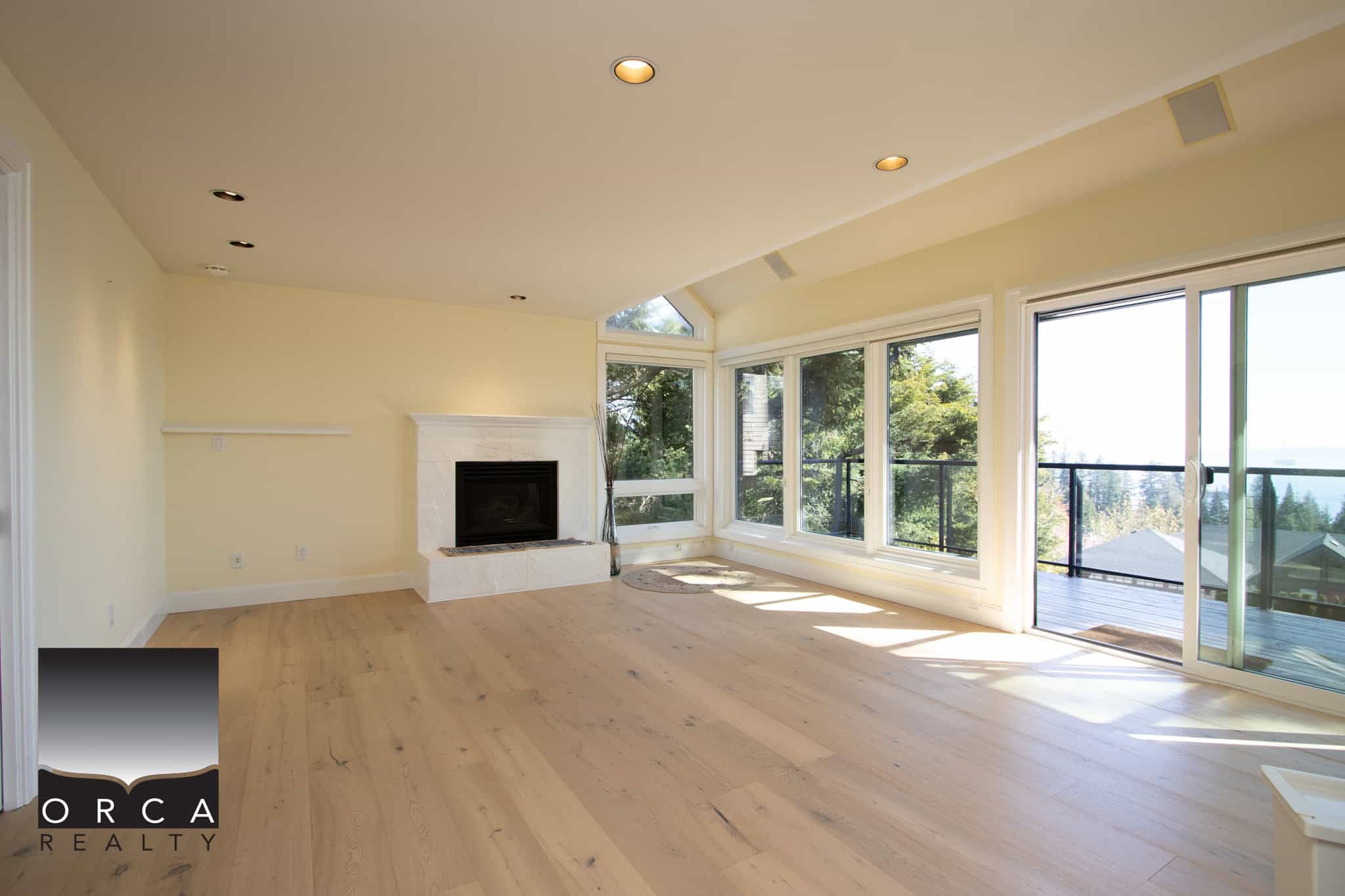 Bright and spacious living room with large windows overlooking lush greenery, showcasing a modern fireplace and hardwood flooring, perfect for real estate listings and property showcase.