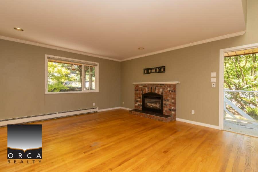 Bright living room with a cozy brick fireplace, hardwood floors, large window, and access to outdoor deck - ideal for Vancouver Island homebuyers.